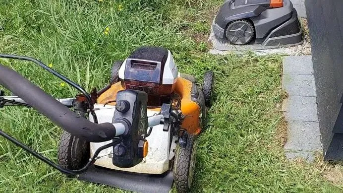 An orange and white battery push mower in long grass, next to a robot mower sitting on its charging dock by a dark wall