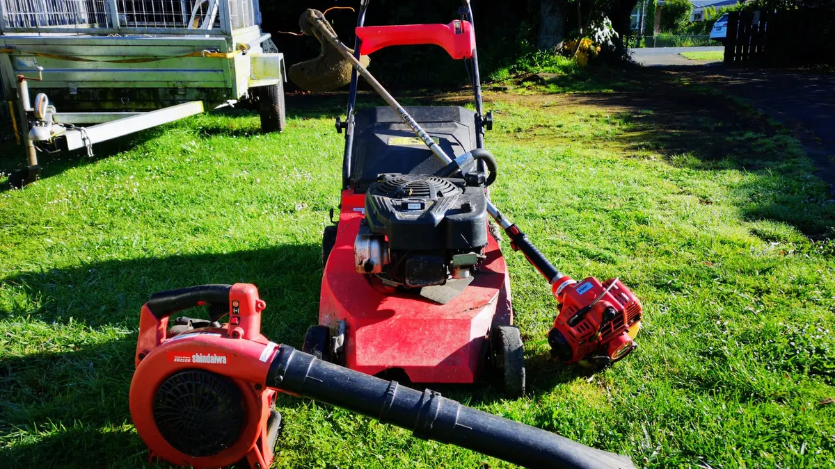 A red push mower, a Shindaiwa blower and a line trimmer lying together on a lawn beside a cage trailer