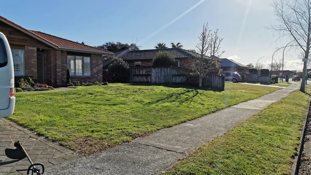 A small mown front lawn and grass berm beside a footpath on a sunny winter morning, with a line trimmer on the paving and the work van just in shot