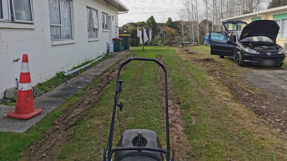 Muddy tyre ruts running down a back lawn beside a house, seen over a mower handle