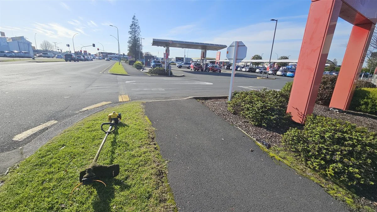 Line trimmer on a grass verge beside a commercial retail forecourt