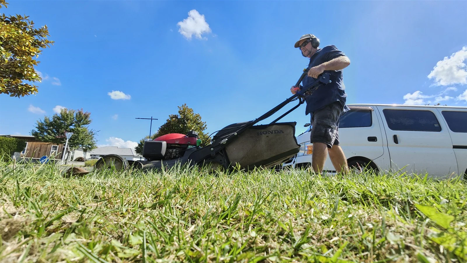 Lawn mowing side hustle: pushing a mower across a small lawn on a sunny afternoon with the vehicle parked at the kerb